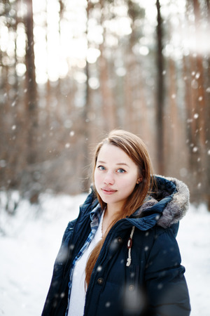 Young woman in winter clothes standing under the snow, looking at camera. Winter forest landscape and snowfall on the background.の写真素材