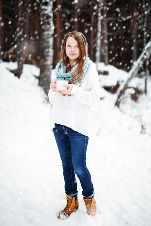 Beautiful woman drinking hot drink in winter park under snowfall. Winter holidays concept. Full height, looking at camera.の写真素材