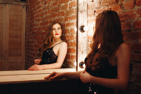 Seductive sexy model posing in loft interior in black dress, looking at vintage mirror with bulbs. Woman with red lips.の写真素材