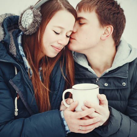 Loving couple in warm clothes drinking hot drink outdoors in winter, man kissing womanの写真素材