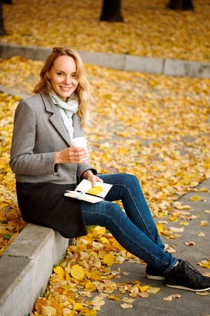 Creative woman with paper notebook and coffee in golden autumn park, looking at camera, full heightの写真素材