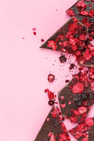 Broken dark chocolate bar pieces with dried red berries and crumbs on pink background