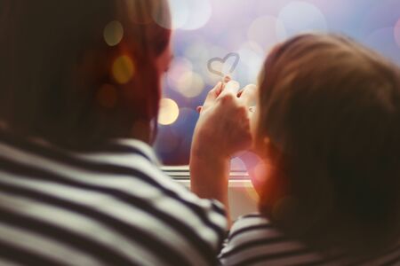 Little girl with her mother drawing heart symbol by fingers on misted window glass with evening lights behind glass. Selective focus.の写真素材