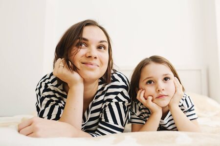 Young beautiful mother and her adorable daughter child lying on bed in bedroom and watching tv or video film attentivly, front view, close-up portraitの写真素材