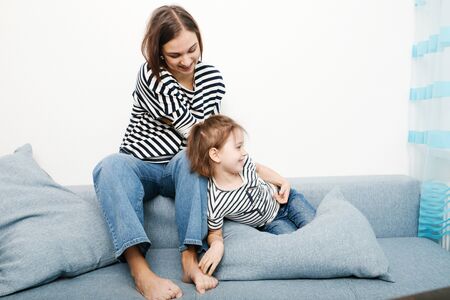 Happy smiling brunette mother with her cute little daughter both in striped t-shirts laughing and having fun together at home in room with white backgroundの写真素材