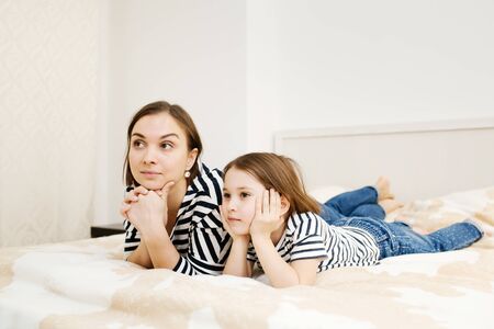 Young beautiful mother and her adorable daughter child lying on bed in bedroom and watching tv or video film attentivly, front view, full heightの写真素材