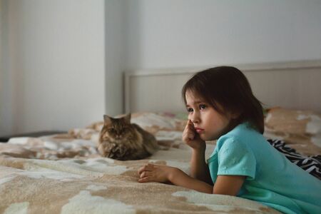 Cute sad little girl watching TV alone in the bedroom with a cat lying on bed, real people, side viewの写真素材