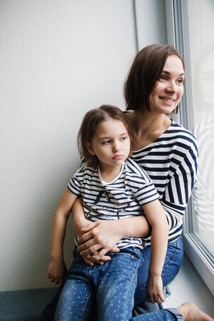 Happy young mother with her upset girl child at home near window, short brunette hair, same clothes, woman smiling and looking out the windowの写真素材