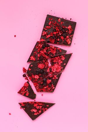 Broken dark chocolate bar pieces with scattered dried red berries on pink background, top view