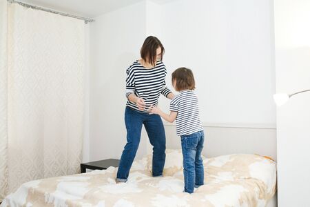 Young happy mother having fun with her cute little daughter, jumping on a bed in bedroom, playing and laughing, active games at home, full body same clothesの写真素材