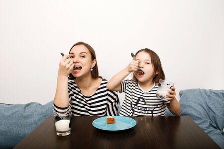 Mother and little kid daughter eating cake together with milk in the morning in light kitchen, having fun and laughing. Same clothes and haircuts.の写真素材