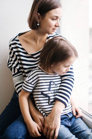 Calm young mother with adorable girl child at home near window, close-up portrait, short brunette hair, same clothes, woman looking in windowの写真素材