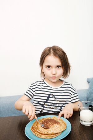 Cute little child eating homemade pancakes with chocolate cream and milk at home kitchen, close-up, girl looking at cameraの写真素材