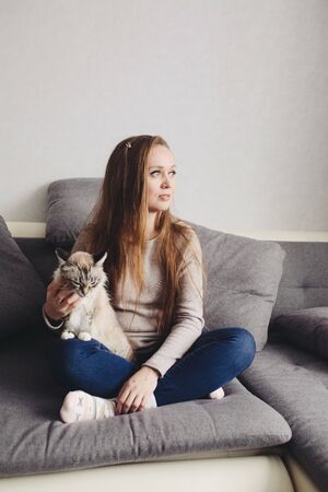 Young trendy woman sitting on the couch at home with domestic cat. Social distance, isolation at home concept. Full body portrait.の写真素材