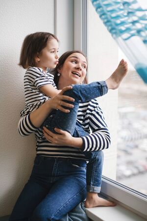Happy young mother having fun with her little girl child at home near window, short brunette hair, same clothes, woman smiling and looking out the windowの写真素材