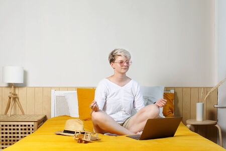 Blonde pretty hipster young woman meditating online sitting on a bed in front of laptop during daybreak, dreaming about travels and adventures の写真素材