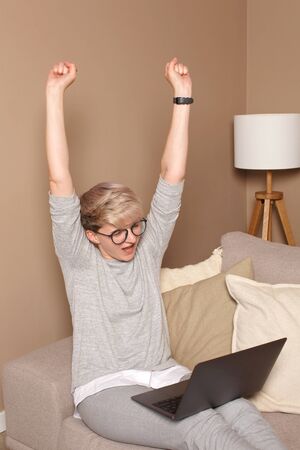 Young excited female celebrating her achievments and wins with her hands up, looking in notebook. Pretty intelligent woman about 30s in casual clothes and glasses sitting on a sofa indoors, working from home.の写真素材