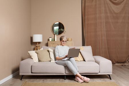 Young female freelancer working or learning from home during self-isolation. Pretty intelligent woman in casual clothes sitting on a sofa typing on a laptop front view.の写真素材