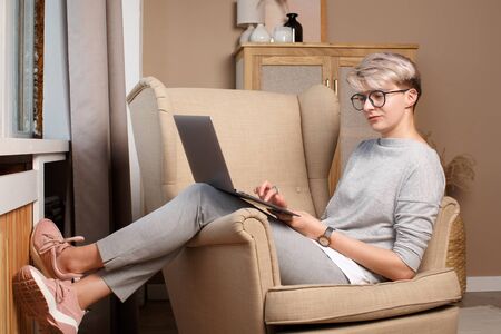Young hiptser female freelancer working from home during self-isolation. Full body portrait of pretty smart woman about 30s in casual clothes sitting in a chair writing in laptop.の写真素材