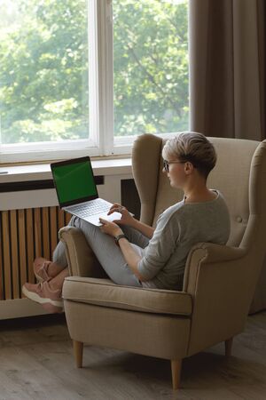 Young hiptser female freelancer working from home during self-isolation. Full body portrait of pretty smart woman about 30s in casual clothes sitting in a chair writing in laptop (green screen mockup).の写真素材