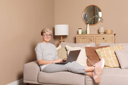 Young happy female freelancer working or learning from home during self-isolation. Full body portrait of pretty intelligent woman about 30s in casual clothes and glasses sitting on a sofa indoors typing on a laptop looking away smiling.の写真素材