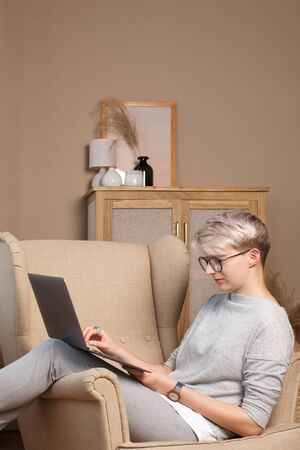 Young hiptser female freelancer working from home during self-isolation. Pretty smart woman about 30s in casual clothes sitting in a chair typing in laptop, side view.の写真素材