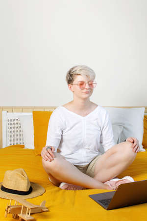 Blonde pretty hipster young woman meditating online sitting on a bed in front of laptop during daybreak, dreaming about travels and adventuresの写真素材