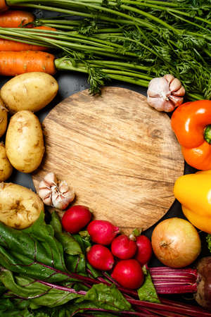 Frame made of fresh assorted vegetables around wooden round cutting board close up: peppers, potatoes, radish, carrotsの写真素材
