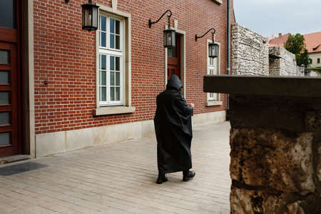 Unrecognizable mysterious man in dark black raincoat walking along red brick wall in old european town, back viewの写真素材