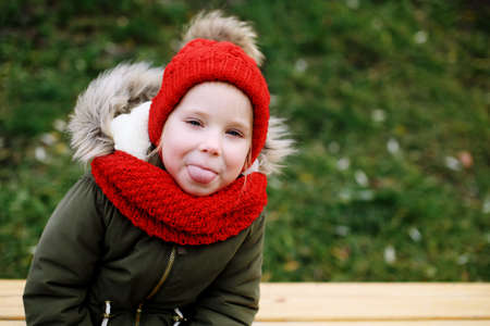 Cute little funny girl kid about 5 years in red knitted hat and scarf having fun showing tongue looking at cameraの写真素材