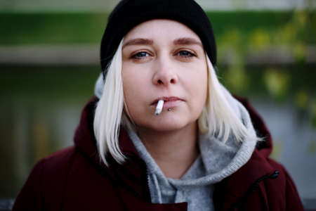 Young hipster millennial woman with lip piercing smoking a cigarette outdoors, looking at camera, headshot close upの写真素材
