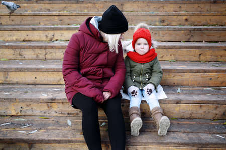 Young mother talking to her cute little daughter outdoors sitting together in warm clothes on wooden stairsの写真素材