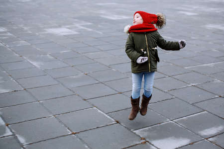 Cute little kid girl in warm outerwear having fun on the street, jumping outdoors, full body portraitの写真素材