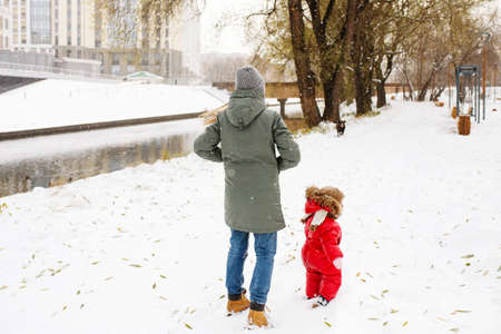 Unrecognizable young woman in winter casual outfit walking with her toddler kid in snowy park near rver, full heightの写真素材