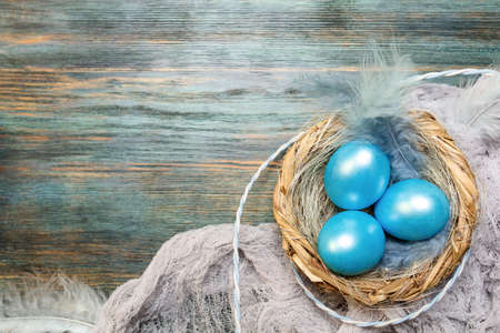 Blue easter background with colored eggs in the nest with feathers on wooden tray, top view with spaceの写真素材