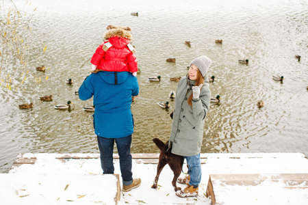 Full height family portrait with one kid and dog in winter casual outfit posing outdoors near river with duck birds, weekend activities and fresh airの写真素材