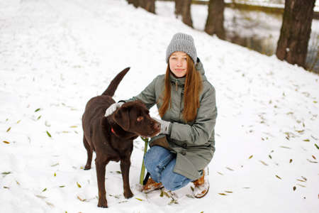 Smiling young woman with brown dog labrador retriever in winter warm outfit walking outdoors in snowy park, full body looking at cameraの写真素材