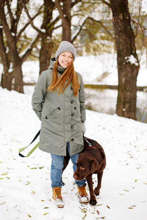 Smiling young woman with brown dog labrador retriever in winter warm outfit walking outdoors in snowy park, full body looking at cameraの写真素材