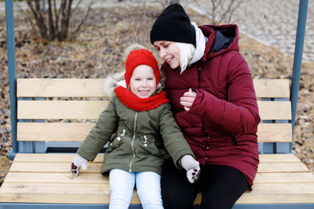 Young mother talking to her cute little daughter outdoors sitting together in warm clothes on wooden swing benchの写真素材