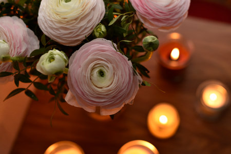 Romantic still life with soft pink ranunculus flowers in a glass vase surrounded by glowing candles....
