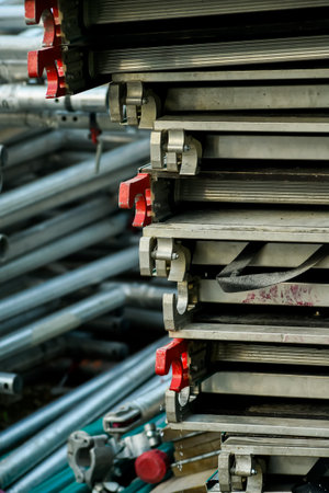 A vertical shot displays neatly stacked aluminum scaffolding components with red latches, alongside a blurred background of metal pipes. This illustrates organized construction equipment storage.の写真素材