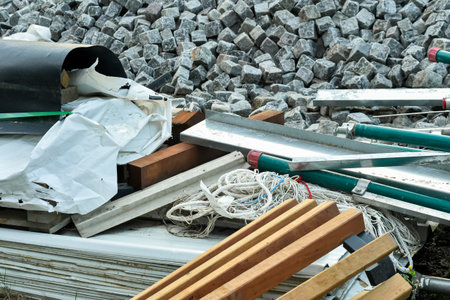 A cluttered view of a construction site showing mixed materials: granite cobblestones, rolled waterproofing membrane, wood planks, metal profiles, and tangled ropes, representing site cleanup.の写真素材