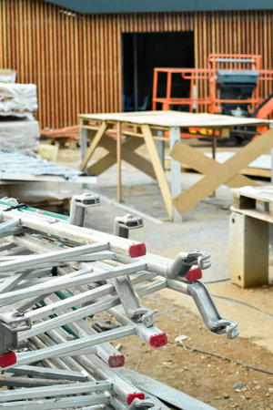 A vertical shot of a construction site with stacked aluminum scaffolding components in the foreground. In the background, wooden walls, sawhorse tables, and other building equipment are visible.の写真素材