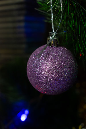 A beautiful, sparkling purple glitter ball ornament hangs from an artificial Christmas tree branch against a dark, blurred background with subtle bokeh lights. Holiday magic and elegance. Vertical.の写真素材