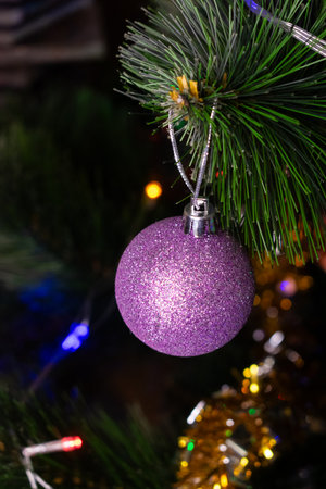 Vertical macro shot of a single purple glitter Christmas bauble hanging on an artificial Pinus branch. Soft, blurred garland lights create a vibrant bokeh effect. Copy space, vertical photo.の写真素材