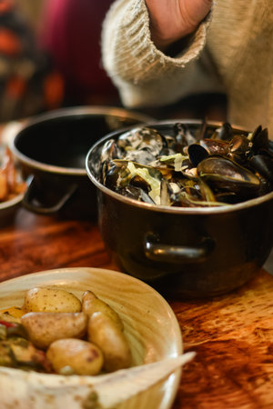 A vertical close-up of a pot filled with freshly cooked mussels, next to a shallow bowl of roasted potatoes and vegetables. Focuses on the texture of the shellfish and rustic meal presentation.の写真素材