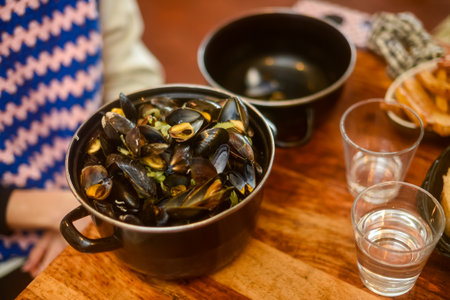 A pot of hot, freshly cooked mussels in a sauce sits on a wooden table, next to two glasses of water or wine. Represents traditional French cuisine and a warm, shared dining experience. Horizontal.の写真素材