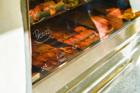 A view into a brightly lit glass display case of a traditional Maltese bakery, showcasing various types of freshly baked, savory Pastizzi. Handwritten labels indicate fillings like Ricotta Cheese.の写真素材