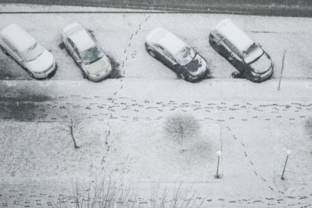 An aerial view of a parking lot in Kyiv, Ukraine, with four cars covered in fresh snow. Numerous footprints crisscross the snowy ground, illustrating challenging winter weather conditions. Horizontal.の写真素材