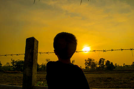 The boy stood waiting for something behind a barbed wire fence in the garden center field , evening light orange sun in beautiful Thailand.のeditorial素材
